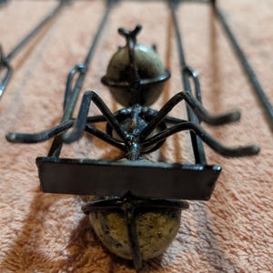 May include: A metal sculpture of an ant carrying a rectangular sign. The ant has a black body and legs, with a spherical head and abdomen. The sign is also black. The sculpture is set against a peach-colored background.