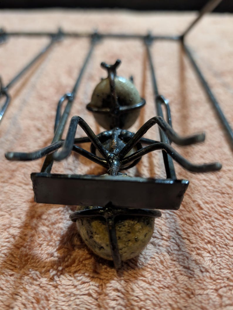 May include: A metal sculpture of an ant carrying a rectangular sign. The ant has a black body and legs, with a spherical head and abdomen. The sign is also black. The sculpture is set against a peach-colored background.