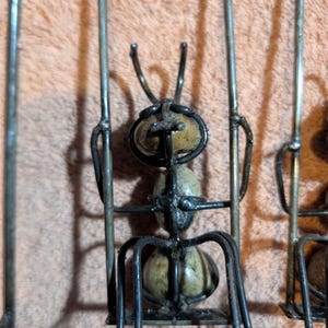 May include: Metal ant sculpture with a detailed design. The ant has a segmented body and legs, with a textured, light-colored body. The sculpture is made of dark metal and is set against a peach-colored background.