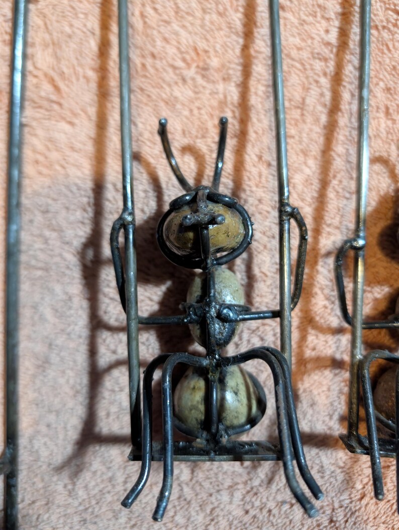 May include: Metal ant sculpture with a detailed design. The ant has a segmented body and legs, with a textured, light-colored body. The sculpture is made of dark metal and is set against a peach-colored background.