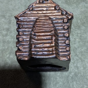 May include: A bronze-coloured decorative cabin figurine. The miniature cabin features a log cabin design with a textured roof and walls. The figurine has a dark base and is made of a metallic material.