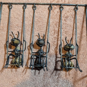 May include: A metal sculpture of three ants on swings. The ants are made from metal and stone, with a dark, aged finish. The swings hang from a horizontal bar, forming a quirky decorative item. The piece is suitable for indoor display.