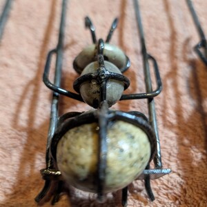 May include: Close-up of a handmade metal and stone sculpture. The sculpture features a cage-like structure with three spherical stones. The metal is dark, and the stones are a speckled beige color. The sculpture is set against a peach-colored background.