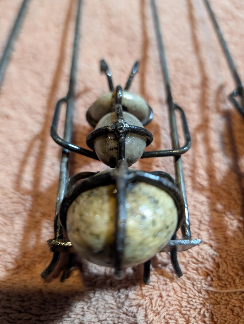 May include: Close-up of a handmade metal and stone sculpture. The sculpture features a cage-like structure with three spherical stones. The metal is dark, and the stones are a speckled beige color. The sculpture is set against a peach-colored background.