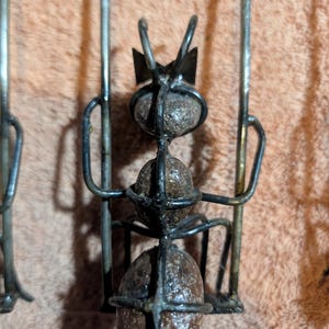 May include: Close-up of a metal ant sculpture with three spherical body segments. The ant has a crown-like structure on its head and is made of dark metal. The sculpture is set against a peach-colored background.