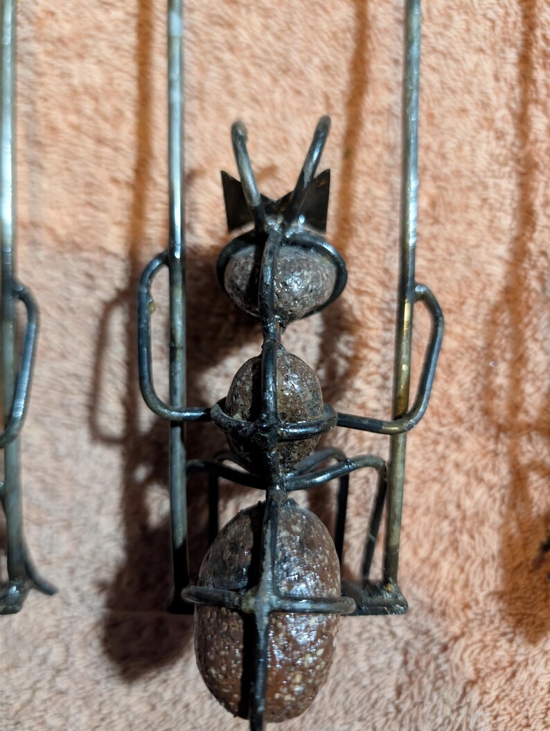 May include: Close-up of a metal ant sculpture with three spherical body segments. The ant has a crown-like structure on its head and is made of dark metal. The sculpture is set against a peach-colored background.