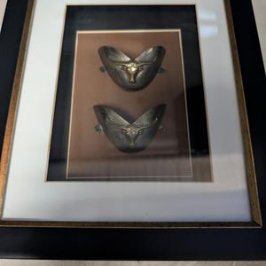 May include: Framed artwork featuring two silver-toned metal sculptures with gold-colored bull heads. The sculptures are mounted on a brown background within a white and black frame with a gold accent.