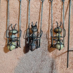 May include: A metal wall hanging featuring three stylized ants on swings. The ants are crafted from stones and metal, with the swings suspended from a horizontal bar. The ants have detailed antennae and are in a playful arrangement.