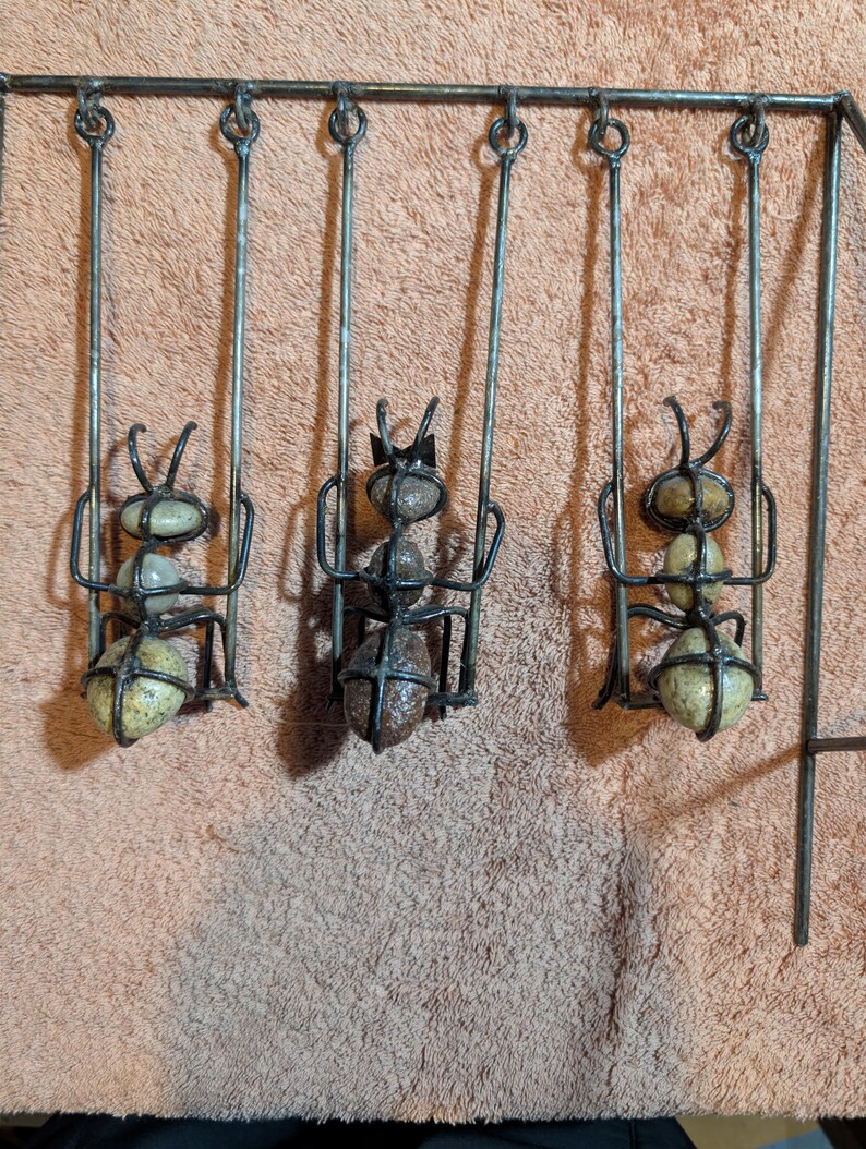 May include: A metal wall hanging featuring three stylized ants on swings. The ants are crafted from stones and metal, with the swings suspended from a horizontal bar. The ants have detailed antennae and are in a playful arrangement.