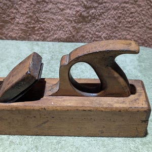 May include: A vintage wooden hand plane with a dark, aged appearance. The plane features a curved handle with a circular opening and a metal blade. The wood has visible grain and signs of wear, suggesting its use in woodworking.