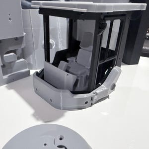 May include: A grey 3D-printed model of a construction vehicle cab. The cab has a detailed interior with a seat and windows. Other grey parts are visible, including a base and side panels. The model is on a white surface.