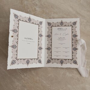 May include: Open wedding invitation with ornate, beige and black border design. The invitation is open, revealing text in Arabic and English. A white ribbon is tied to the side.