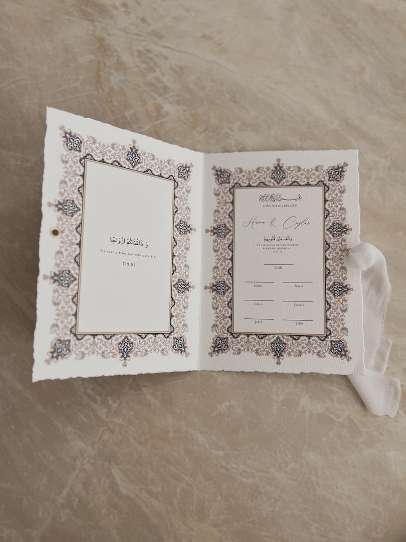 May include: Open wedding invitation with ornate, beige and black border design. The invitation is open, revealing text in Arabic and English. A white ribbon is tied to the side.