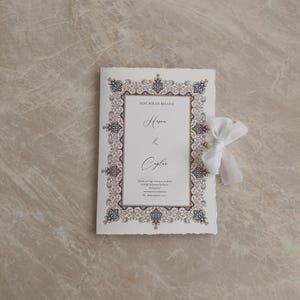 May include: A cream-colored wedding invitation with ornate, dark brown and gold border design. The invitation features the names "Hesna & Ceylan" in elegant script, with additional text below. A white ribbon is tied to the side.