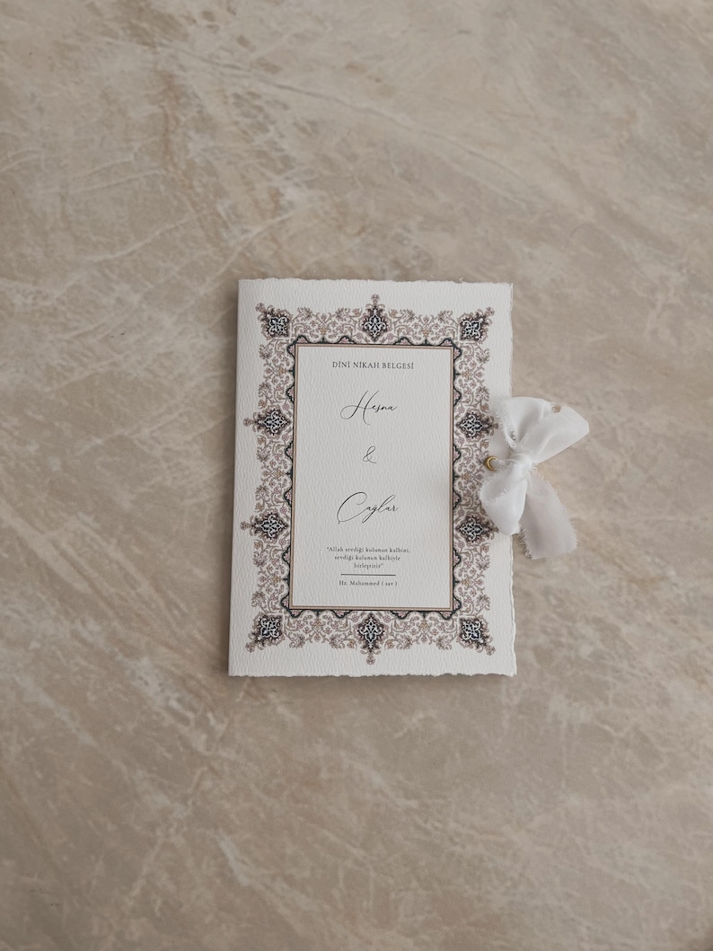 May include: A cream-colored wedding invitation with ornate, dark brown and gold border design. The invitation features the names "Hesna & Ceylan" in elegant script, with additional text below. A white ribbon is tied to the side.