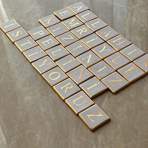 Könnte beinhalten: Anordnung quadratischer Kacheln mit goldfarbenen Buchstaben auf hellgrauem Hintergrund. Die Kacheln buchstabieren eine türkische Nachricht. Jede Kachel hat einen goldenen Rand. Die Kacheln sind auf einer neutral getönten Oberfläche angeordnet.