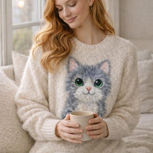 May include: Cream-colored sweater featuring a large, detailed illustration of a gray cat with big green eyes. The sweater is worn by a person holding a beige mug. The overall style is cozy and casual.