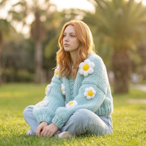 May include: A pale blue knitted sweater decorated with large daisy appliqués. The sweater is worn with blue jeans. The person is sitting on green grass in a park, with palm trees in the background.