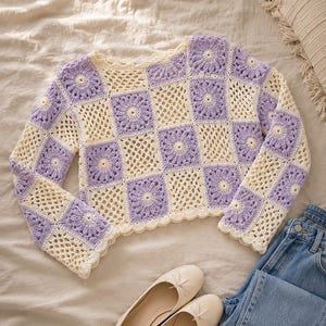 May include: A cream and lavender crochet sweater with a floral square pattern. The long-sleeved top is paired with light-colored flats and blue jeans. The sweater's design features a scalloped edge.