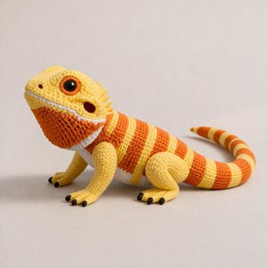 Puede incluir: Un dragón barbudo de ganchillo en amarillo, naranja y blanco. El reptil tiene garras negras y una cabeza detallada con una prominente barba naranja. El cuerpo presenta rayas alternas naranjas y amarillas.