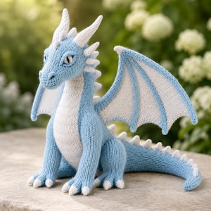 May include: A hand-knitted dragon plush toy featuring a light blue body with a white underbelly, white wings, and white spikes running along its back and tail. The creature has pointed ears, a long tail, and detailed stitching throughout its form.