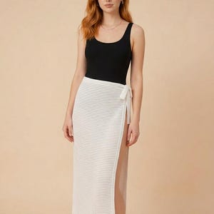 Op de afbeelding: Een vrouw draagt een zwarte tanktop en een witte wikkelrok met open weefsel. De rok heeft een hoge split en wordt met een wit lint in de taille geknoopt. De outfit is eenvoudig en elegant.