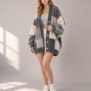 Puede incluir: Un cárdigan de gran tamaño con diseño de patchwork. El suéter presenta grandes cuadrados grises, blanquecinos y carbón. Tiene una botonadura y mangas largas. La modelo lleva zapatillas blancas y pantalones cortos. La longitud es de unos 76 cm.