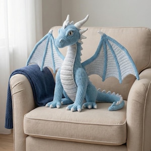 Puede incluir: Un gran dragón tejido a ganchillo en azul claro y blanco está sentado en un sillón beige. El dragón tiene grandes alas, cuernos y una larga cola. Una manta azul está sobre el brazo de la silla.