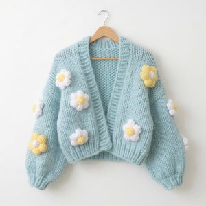 May include: A light blue, open-front cardigan sweater with a chunky knit design. The sweater is adorned with white and yellow daisy appliqués. The garment is hanging on a wooden hanger against a white background.