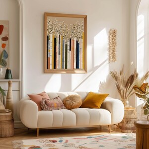 Modern poster of painted book spines and daisy blooms in beige, mustard, and navy tones, hanging above a cream sofa in a bright living room.