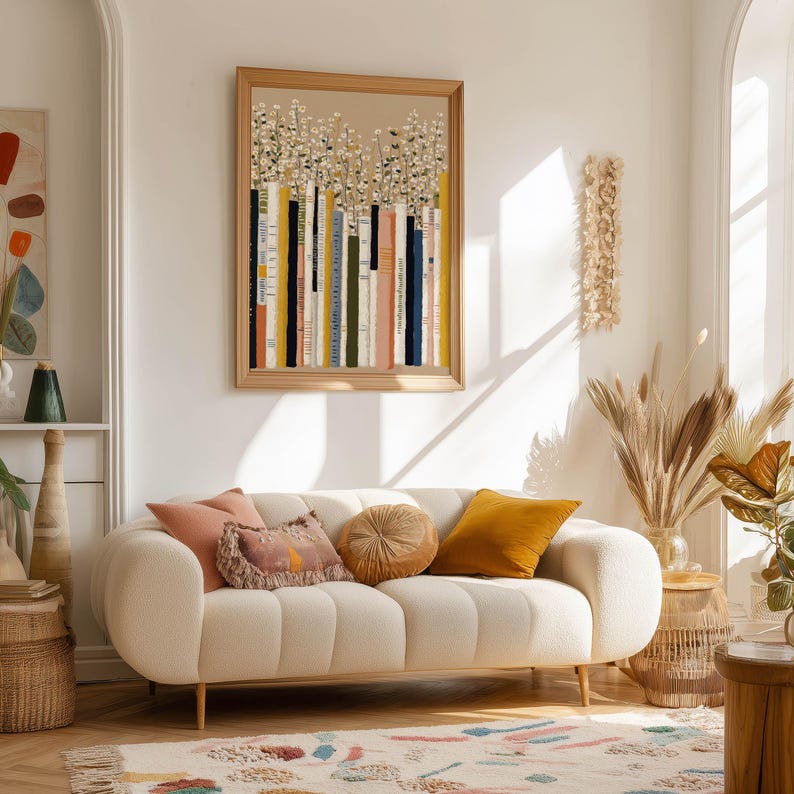 Modern poster of painted book spines and daisy blooms in beige, mustard, and navy tones, hanging above a cream sofa in a bright living room.
