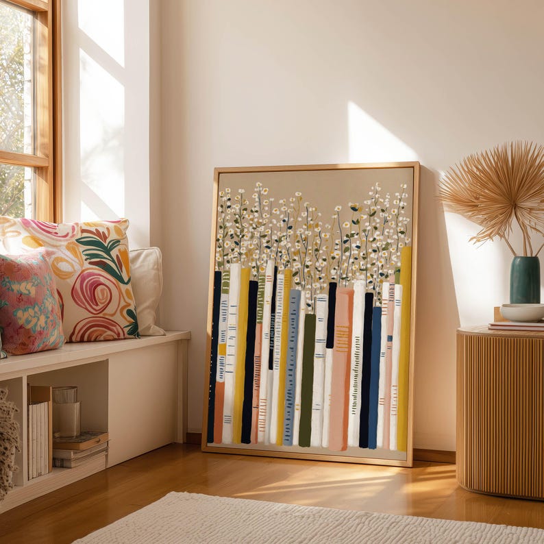 Playful bookshelf art print with tall books and tiny white flowers in warm neutrals, leaning beside a window seat with pillows, stacked books, and a ribbed cabinet.