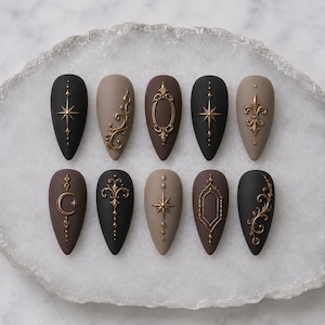 May include: A set of ten almond-shaped press-on nails featuring a matte finish in shades of black, dark brown, and taupe. Each nail is decorated with raised gold-toned metallic embellishments, including starbursts, filigree swirls, and ornate frames.