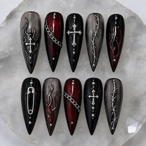May include: A set of ten stiletto-shaped press-on nails featuring a black and deep red color palette. The designs include silver-toned metal embellishments such as chains, crosses, safety pins, barbed wire, and raised flame patterns on glossy surfaces.