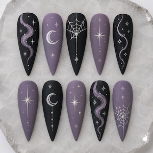 May include: A set of ten almond-shaped press-on nails featuring a matte finish in black and muted purple. The designs include silver hand-painted motifs such as crescent moons, spider webs, swirling smoke-like lines, and various four-pointed stars.