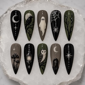 May include: A set of ten almond-shaped press-on nails featuring a dark color palette of black, olive green, and grey. The designs include raised 3D elements such as a silver crescent moon, a black raven, a white ghost, a spider web, bare tree branches, mushrooms, a lantern, an owl, a tombstone with a cross, and a starburst.