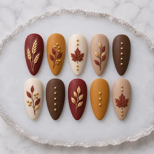May include: A set of ten almond-shaped press-on nails featuring a matte finish in shades of burgundy, mustard yellow, cream, beige, and dark brown. The designs include raised gold metallic wheat stalks, leaves, maple leaves, and small gold studs.