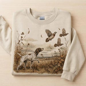 May include: A beige crewneck sweatshirt featuring a detailed graphic print of a spotted hunting dog standing in a field of tall grass, with three birds flying overhead. The Gildan brand label is visible on the inside of the collar.