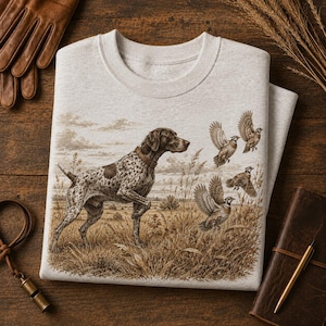 May include: A light heather gray crewneck sweatshirt featuring a detailed graphic print of a brown and white spotted hunting dog standing in a field of tall grass, with three game birds flying in the sky above the landscape.