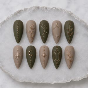 May include: A set of ten almond-shaped press-on nails in matte olive green and beige. The nails feature raised gold-toned metallic accents including delicate leaf vines, thin wavy lines, geometric arches, starbursts, and a crescent moon design.