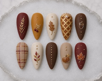 May include: Ten almond-shaped press-on nails featuring autumn-themed designs in shades of burgundy, mustard yellow, cream, and brown. Embellishments include gold metallic wheat, leaves, acorns, pumpkins, plaid patterns, and small gold studs.