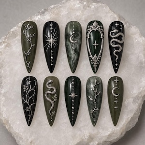 May include: Ten stiletto-shaped press-on nails in shades of black and dark green. The designs feature silver metallic embellishments including snakes, crescent moons, stars, and intricate vine patterns, with some nails displaying a marbled finish.