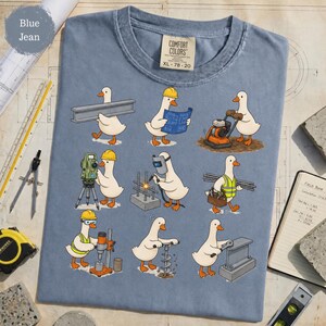 May include: A blue jean colored t-shirt featuring a graphic print of nine white ducks performing various construction tasks, including welding, surveying, and carrying building materials. The shirt has a Comfort Colors label at the neck.