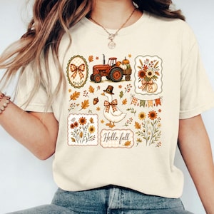 Peut inclure: Un t-shirt couleur crème avec une impression graphique d'illustrations sur le thème de l'automne, incluant un tracteur rouge, une oie portant un chapeau de pèlerin et un nœud à carreaux, des tournesols, des citrouilles, des feuilles et le texte "Hello fall".