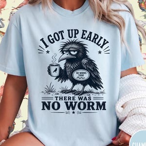 May include: A light blue t-shirt featuring a black graphic of a grumpy crow holding a coffee mug. The text reads I GOT UP EARLY, NO WORMS TODAY, and THERE WAS NO WORM. The design includes stars and grass details, creating a humorous illustration.