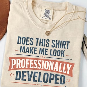 Peut inclure: Un t-shirt beige à manches courtes avec une étiquette Comfort Colors en taille small. Le motif présente un texte bleu foncé indiquant DOES THIS SHIRT MAKE ME LOOK PROFESSIONALLY DEVELOPED, avec une bannière rouge et des icônes.
