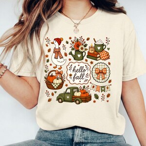 Peut inclure: Un t-shirt couleur crème avec une impression graphique colorée portant le texte "hello fall". Le motif comprend une oie avec une écharpe, un arrosoir avec des fleurs, une tasse sur des livres, un panier de citrouilles, un camion vert et un nœud.