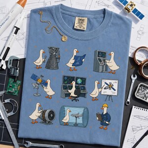 Peut inclure: Un t-shirt bleu à col rond orné d'un imprimé représentant des canards blancs effectuant diverses tâches d'ingénierie aérospatiale, comme travailler sur des moteurs de fusée, des satellites et des consoles, avec l'étiquette Comfort Colors au col.
