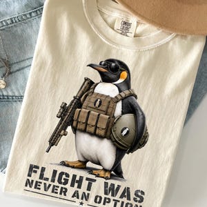 Peut inclure: Un t-shirt couleur crème avec un motif représentant un manchot portant des lunettes de soleil, un gilet tactique, un fusil et un casque. Le texte sous le manchot indique "FLIGHT WAS NEVER AN OPTION". Le t-shirt porte une étiquette Comfort Colors.