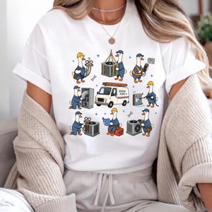 Op de afbeelding: Een wit t-shirt met een grafische print van stripganzen verkleed als HVAC-monteurs. De ganzen voeren verschillende taken uit, zoals het controleren van meters, het tillen van apparatuur en het lezen van blauwdrukken. Een witte bestelwagen met het opschrift GOOSE HVAC staat in het midden.
